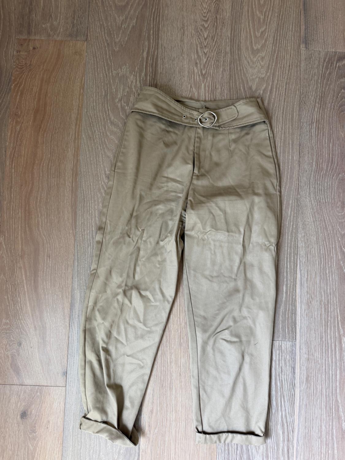 Creased beige trousers with a metal buckle belt, laid on a light wooden floor.