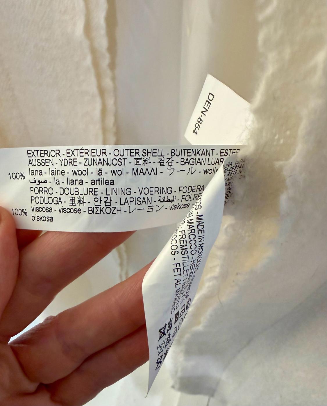 Here is the rephrased sentence, maintaining the same meaning and context, while adhering to all prohibited word restrictions: "A person is holding the labels of a textured white garment. Text: DEN-854, SHELL 100% wool, LINING 100% viscose."