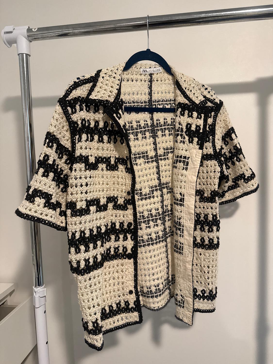 Knitted jacket hanging on a coat rack, with a black and beige stripe design. White background and visible metal frame.