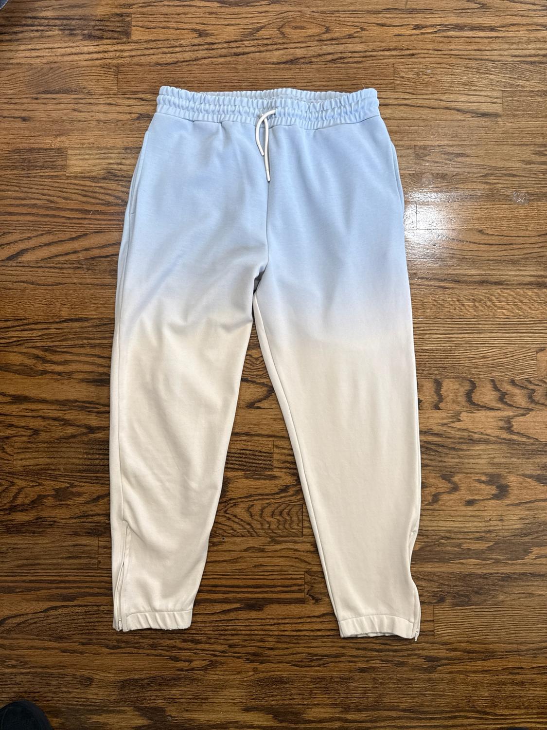 Light blue to cream gradient athletic pants on a wooden floor. Elastic waistband with a white drawstring and zippers at the ankles.