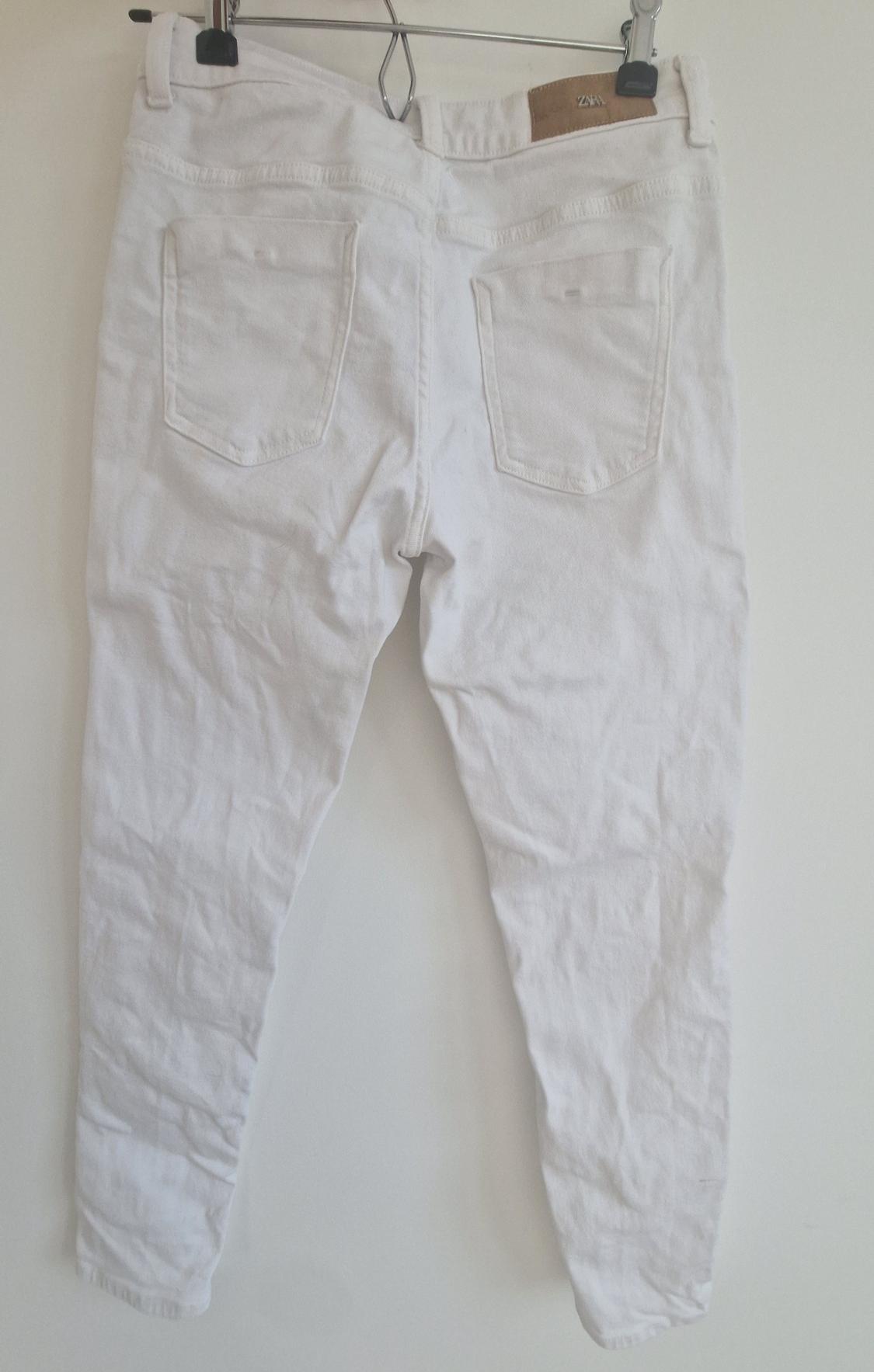 White trousers hanging on a hanger, viewed from behind. They have two back pockets and a brown label on the waistband.