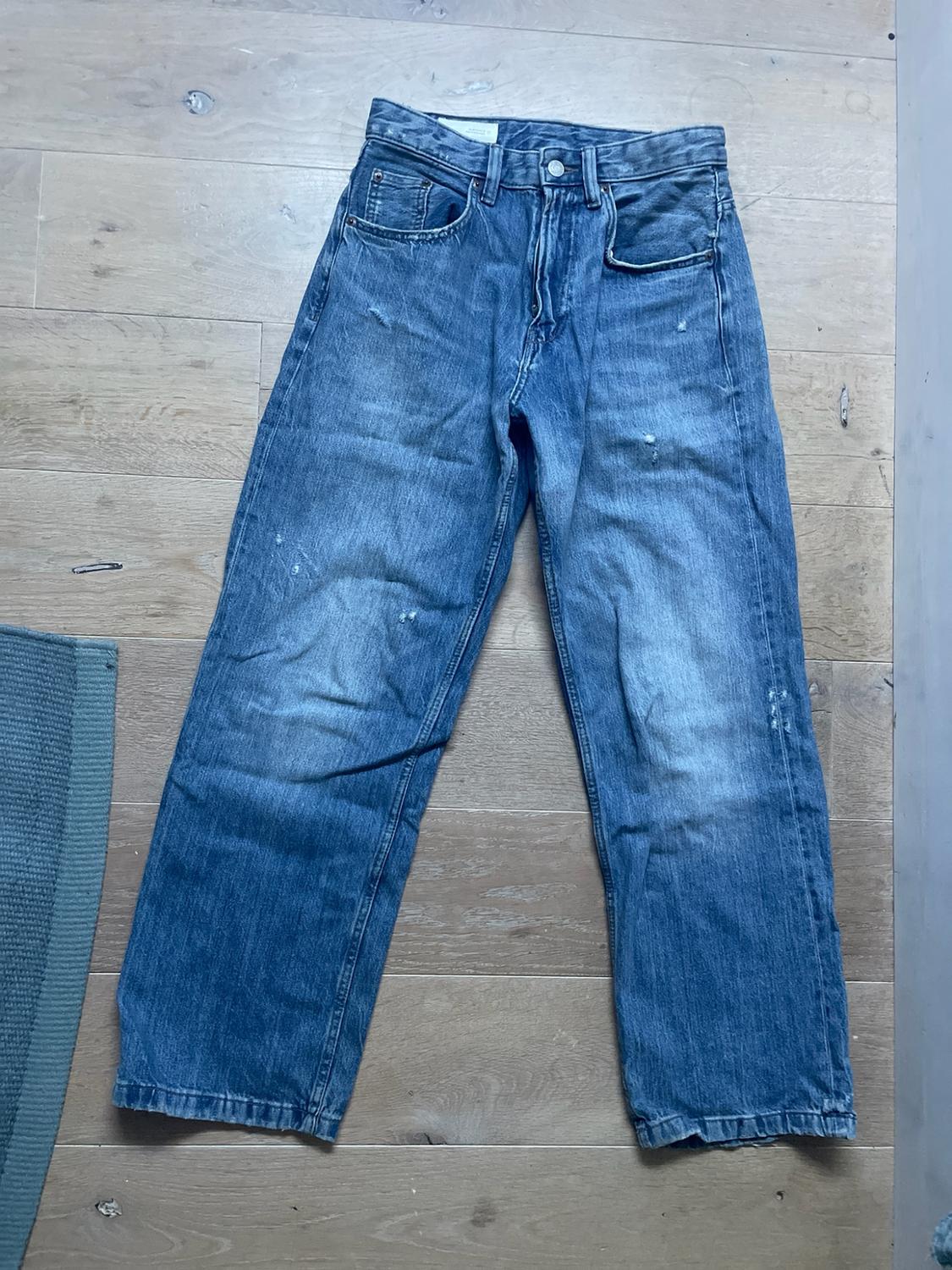 Blue straight-leg jeans with a washed effect and slight distressed detailing, laid out on a light wooden floor.