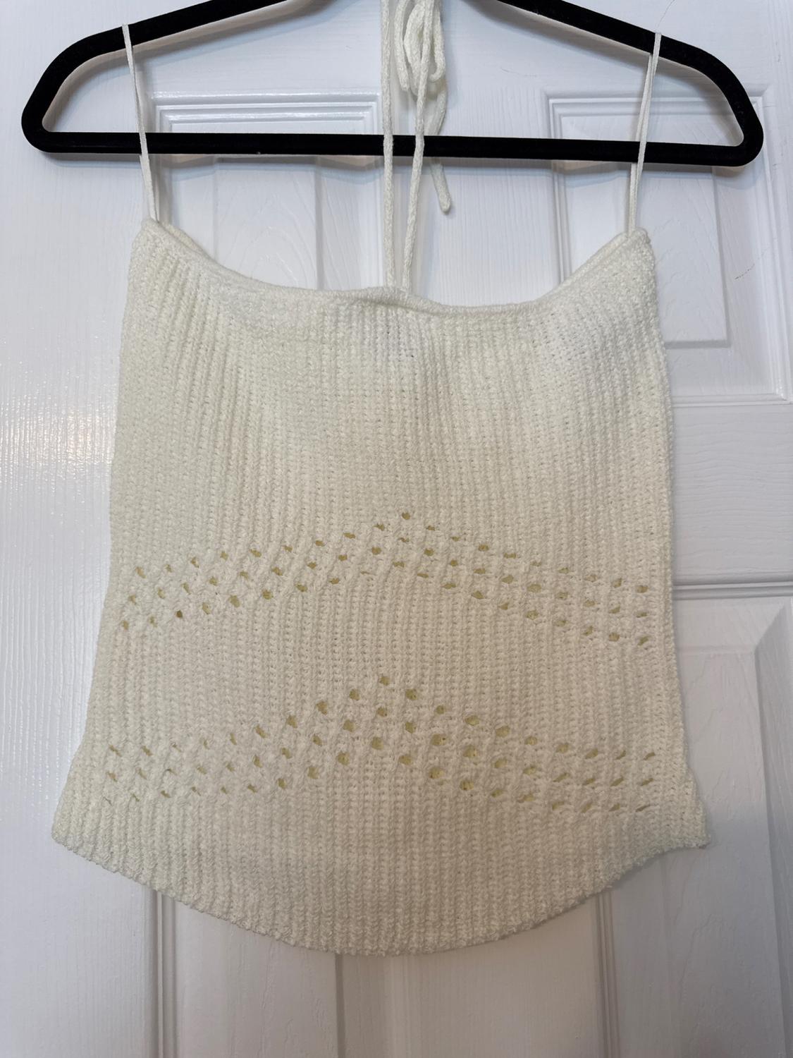 White knitted top hanging on a black hanger over a white door. Design with thin straps and openwork details.