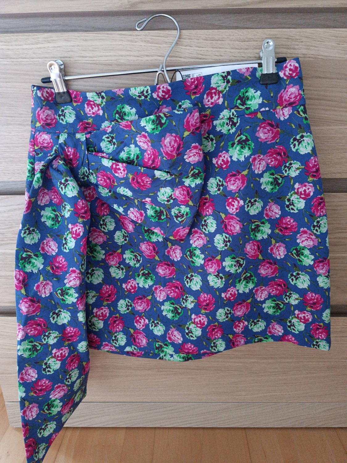Short blue skirt with a pink and green floral print and a large side bow. It is hanging on a metal hanger against a wooden background.