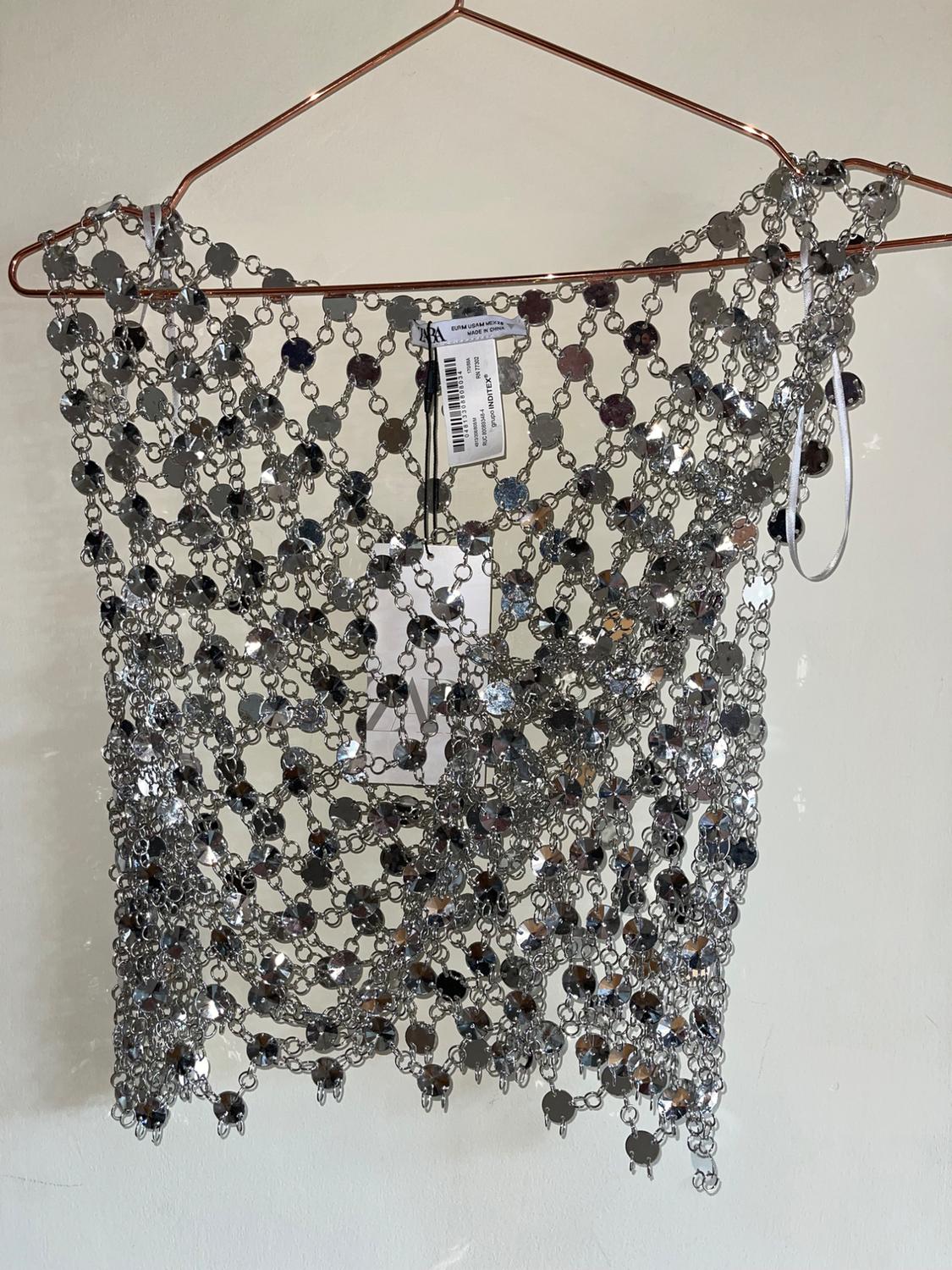 Silver-tone metallic mesh top with sparkly sequins, hanging on a copper metal hanger against a white backdrop. Labels visible.