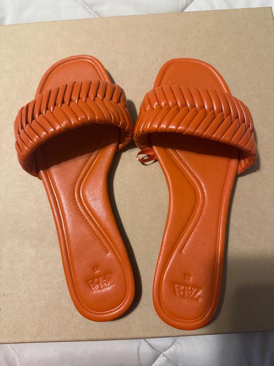 Orange sandals with a braided design on the upper, on a beige background. Visible text: "ZARA".