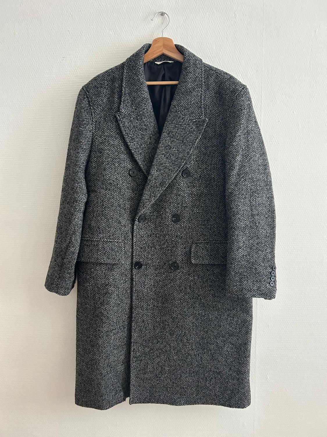Grey wool coat with a herringbone pattern, hanging on a wooden hanger against a white wall. Double-breasted design.