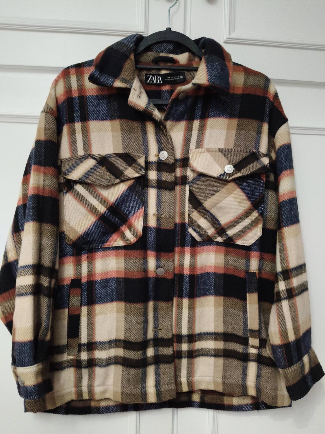 Checked jacket in shades of beige, blue, and red with front pockets and buttons, hanging on a hanger. Visible label: ZARA.