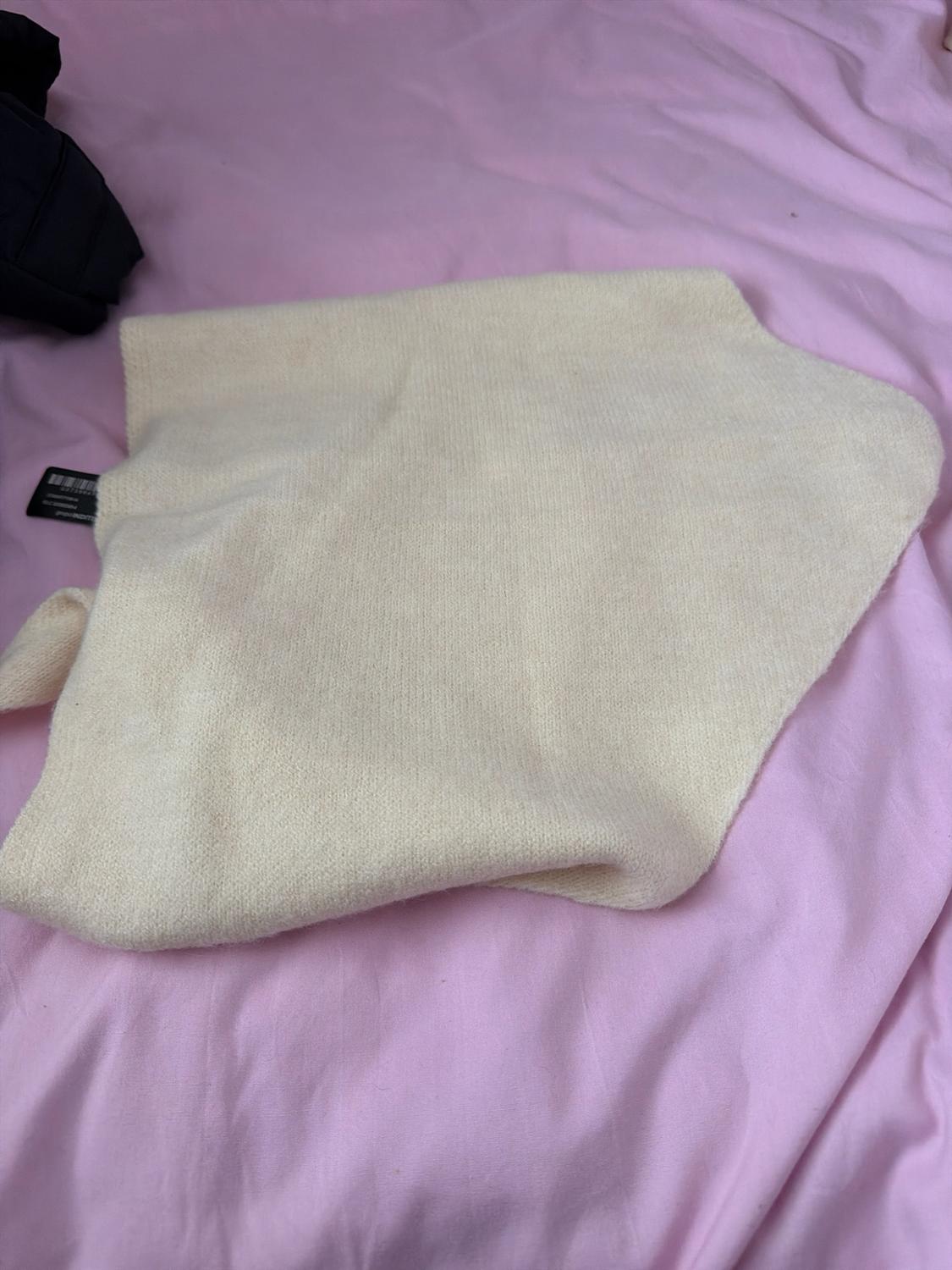 Beige knitted garment folded on a crumpled light pink fabric. A black label with a barcode peeks out from the left edge.