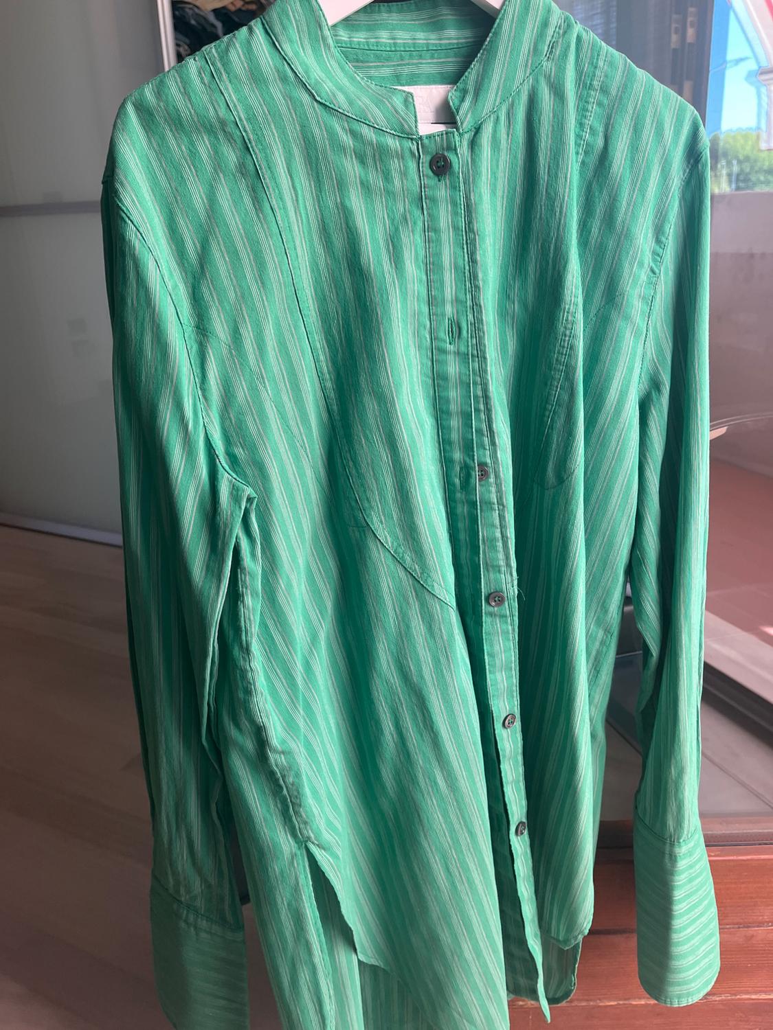 Green long-sleeved shirt with fine white stripes and a mandarin collar, hanging on a hanger in a bright interior with a wooden floor.