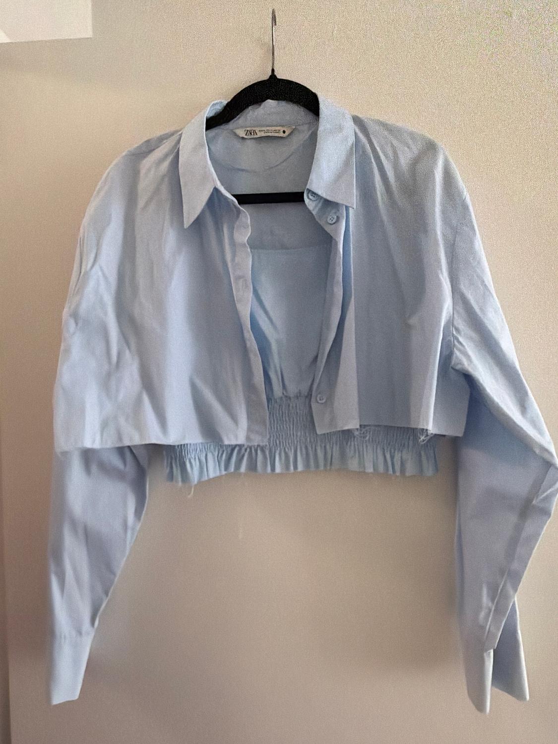 Light blue, long-sleeved, cropped shirt, hanging on a black hanger. Visible label with the text "Zara".