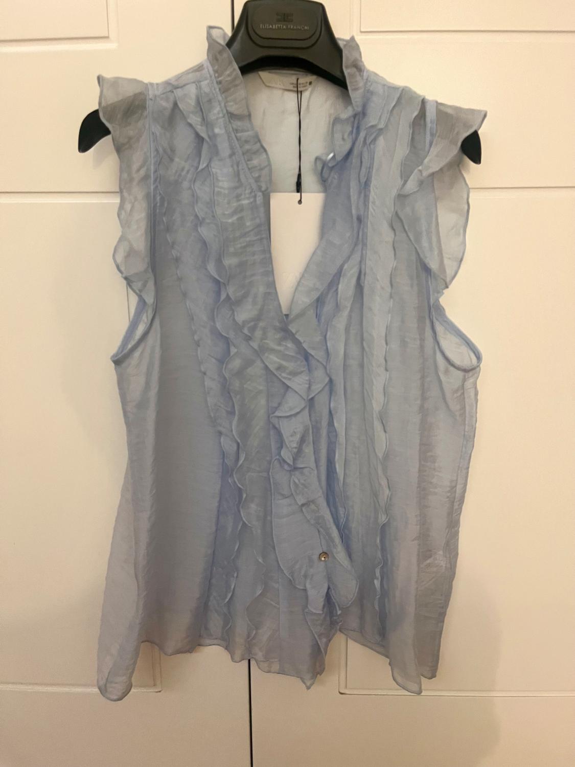 Light blue sleeveless blouse with ruffles, hanging on a black Elisabetta Franchi hanger, on a white background.