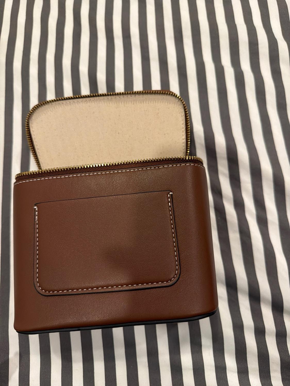 Structured brown bag with an open zipper, light interior, and front pocket with topstitching, set against a black and white striped background.