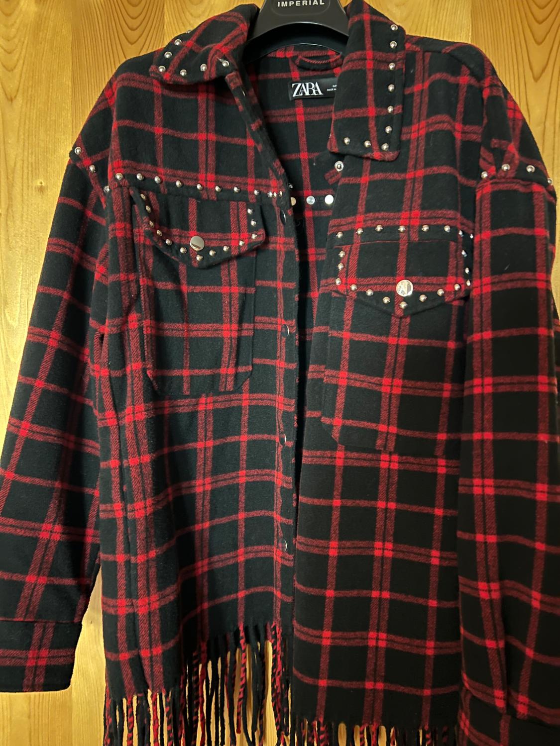 Red and black check overshirt with studs and fringing. Hanging on a hanger with IMPERIAL text on wood. Label with ZARA text.