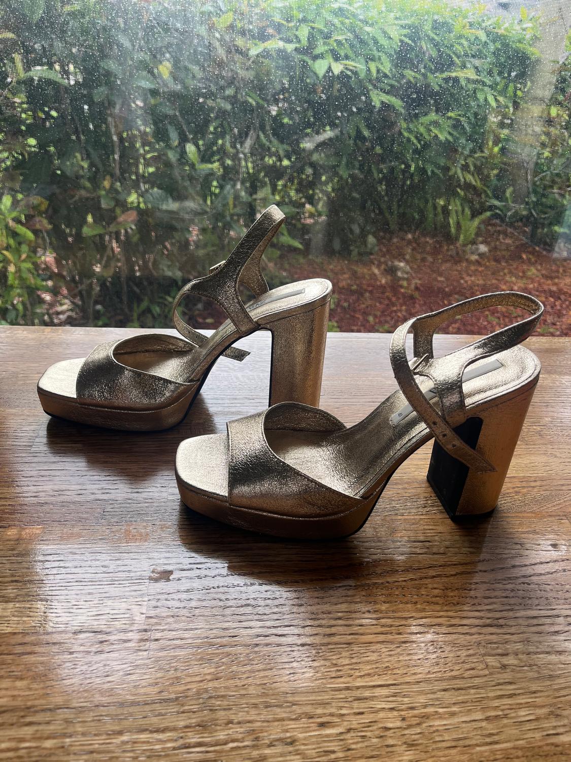 Gold chunky-heeled sandals on a wooden table, with foliage visible through a window in the background.