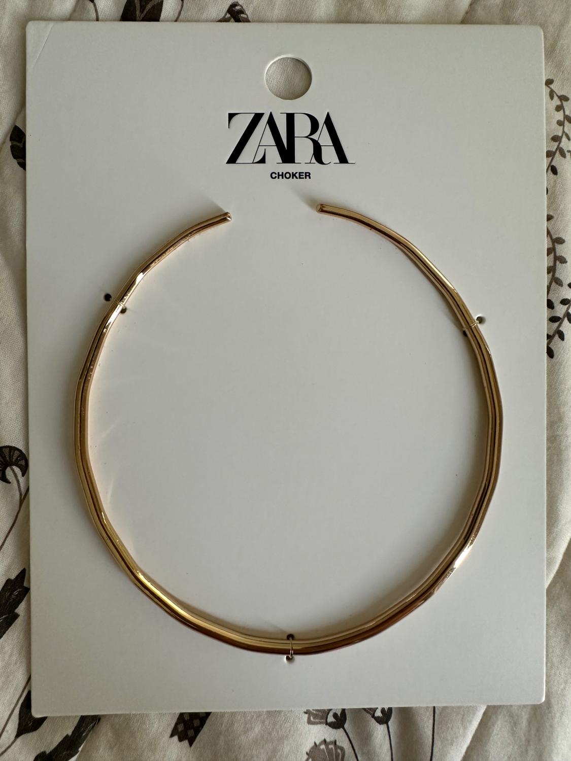 Rigid gold choker attached to a white card bearing the text ZARA CHOKER, against a patterned fabric background.