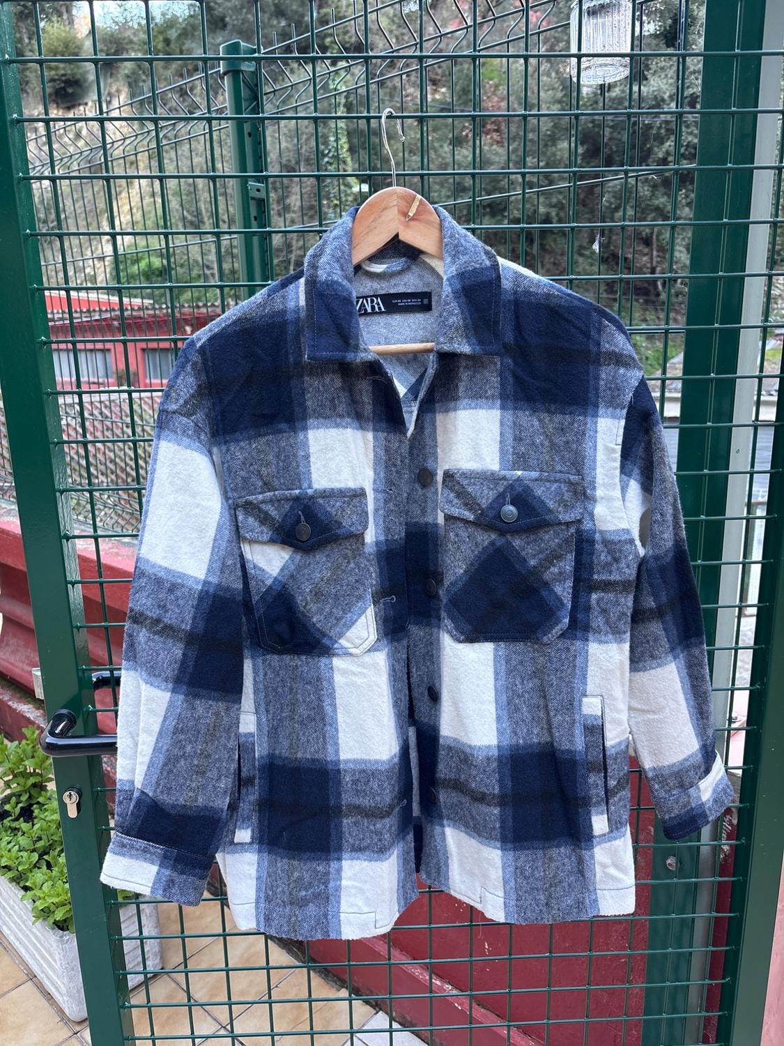 Blue and white checked shirt hanging on a wooden hanger over a green railing. Visible label with text "ZARA".