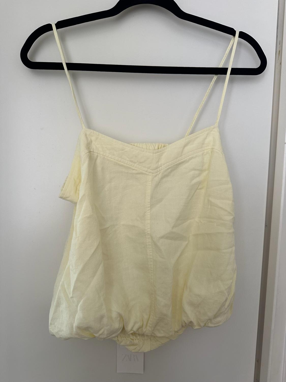 Light yellow camisole top with thin straps and an elasticated hem hanging on a black hanger against a white background. Bottom label with the text ZARA.