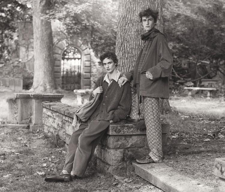 Black and white: Two models in loose-fitting clothes pose in a rustic setting with trees and stone walls, conveying a relaxed vibe.