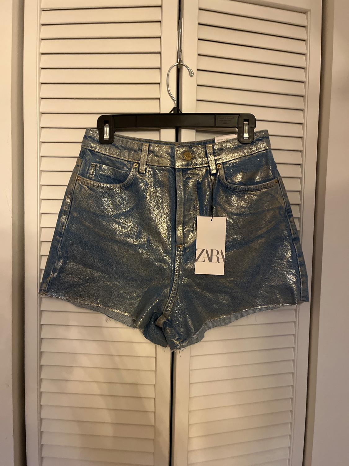 Silver metallic shorts hanging on a black hanger in front of white shutter doors. Visible tag with the text "ZARA".