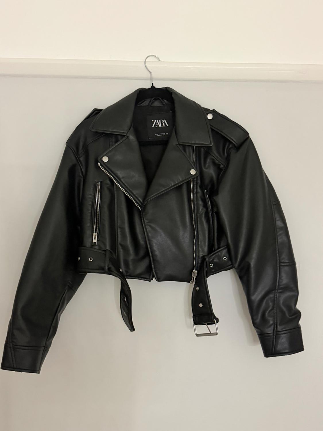 Black biker jacket hanging on a hanger against a white background. Short design with silver zips, a belt and a label bearing the text ZARA.