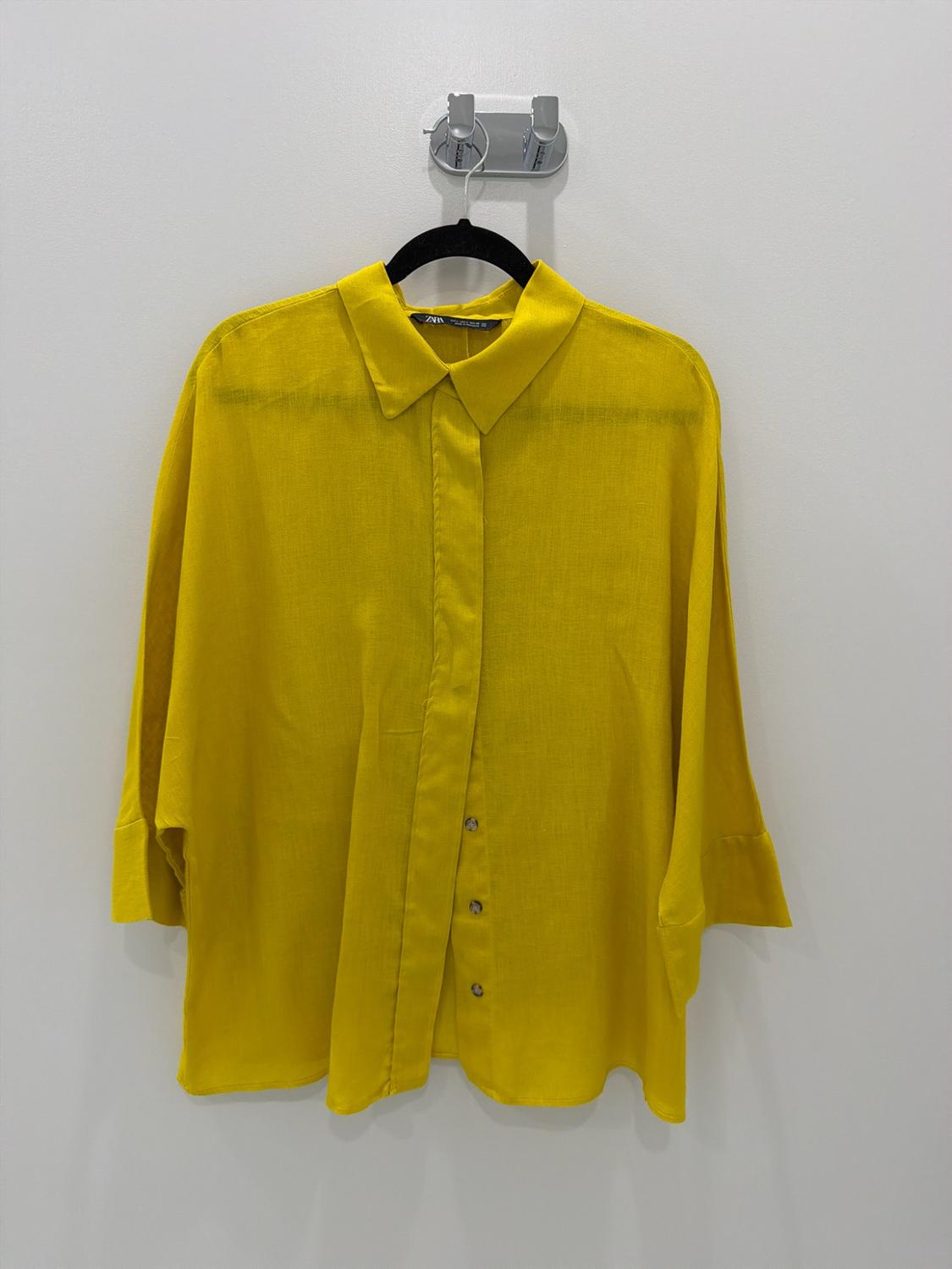 Yellow three-quarter-sleeve shirt hanging on a black hanger against a white wall. Label with ZARA text.