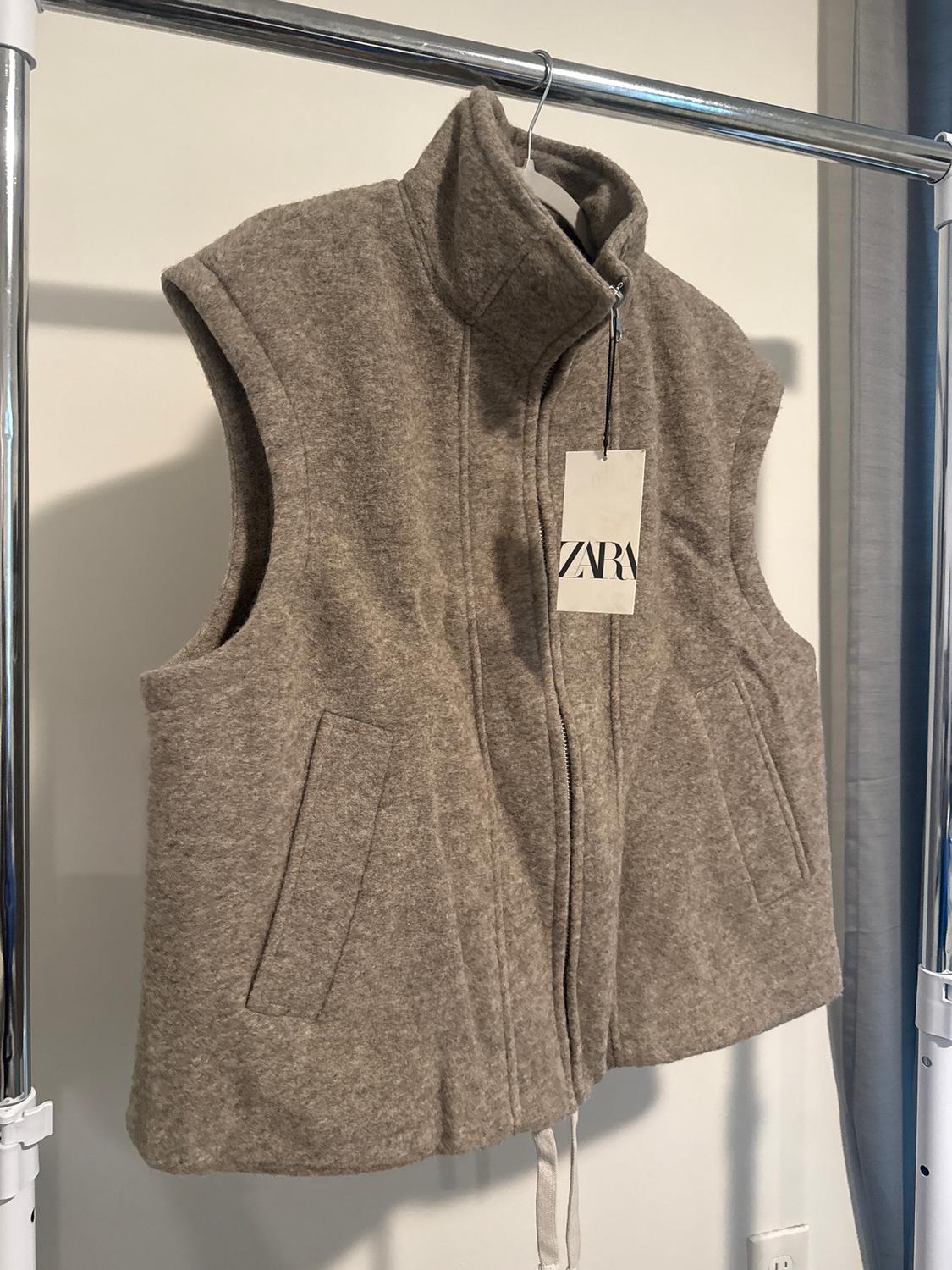 Grey sleeveless waistcoat hanging on a coat hanger, with a 'ZARA' label. White background and grey curtain to the right.