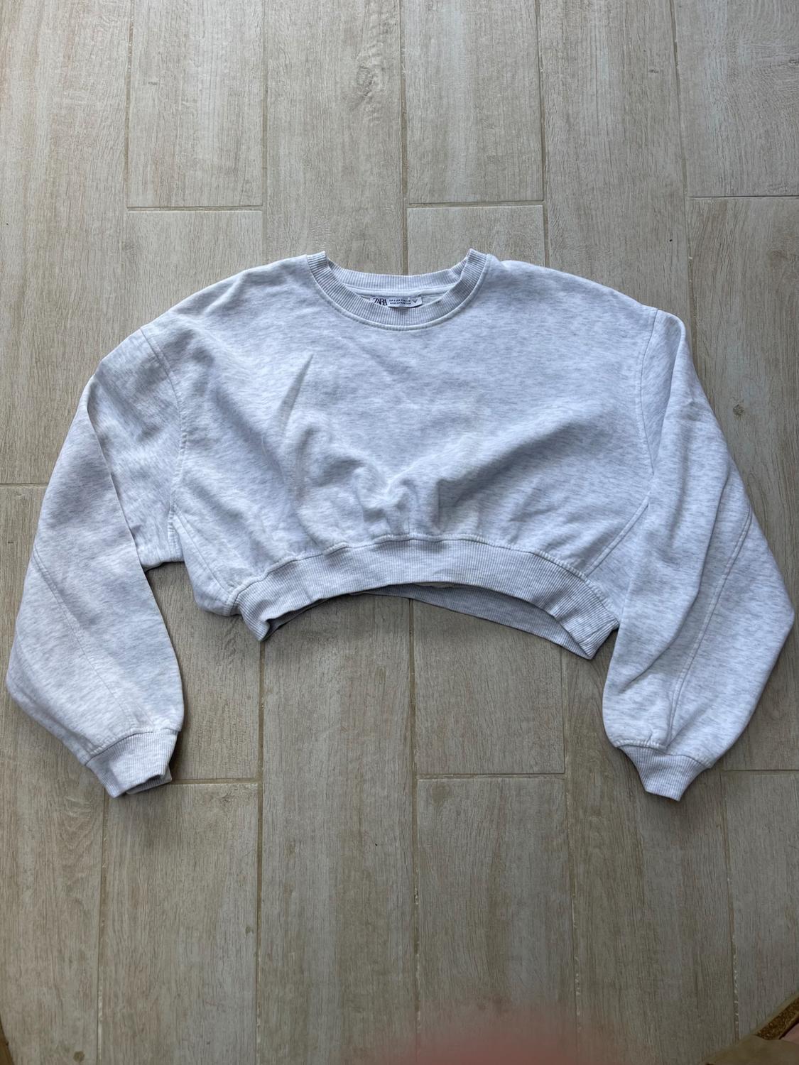 Light grey cropped sweatshirt with a crew neck and long sleeves, on a beige wood floor background.