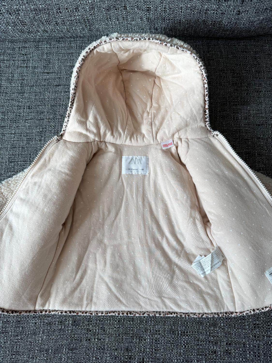 Baby jacket open on a grey sofa, showing the beige inner lining with white dots and visible tags.