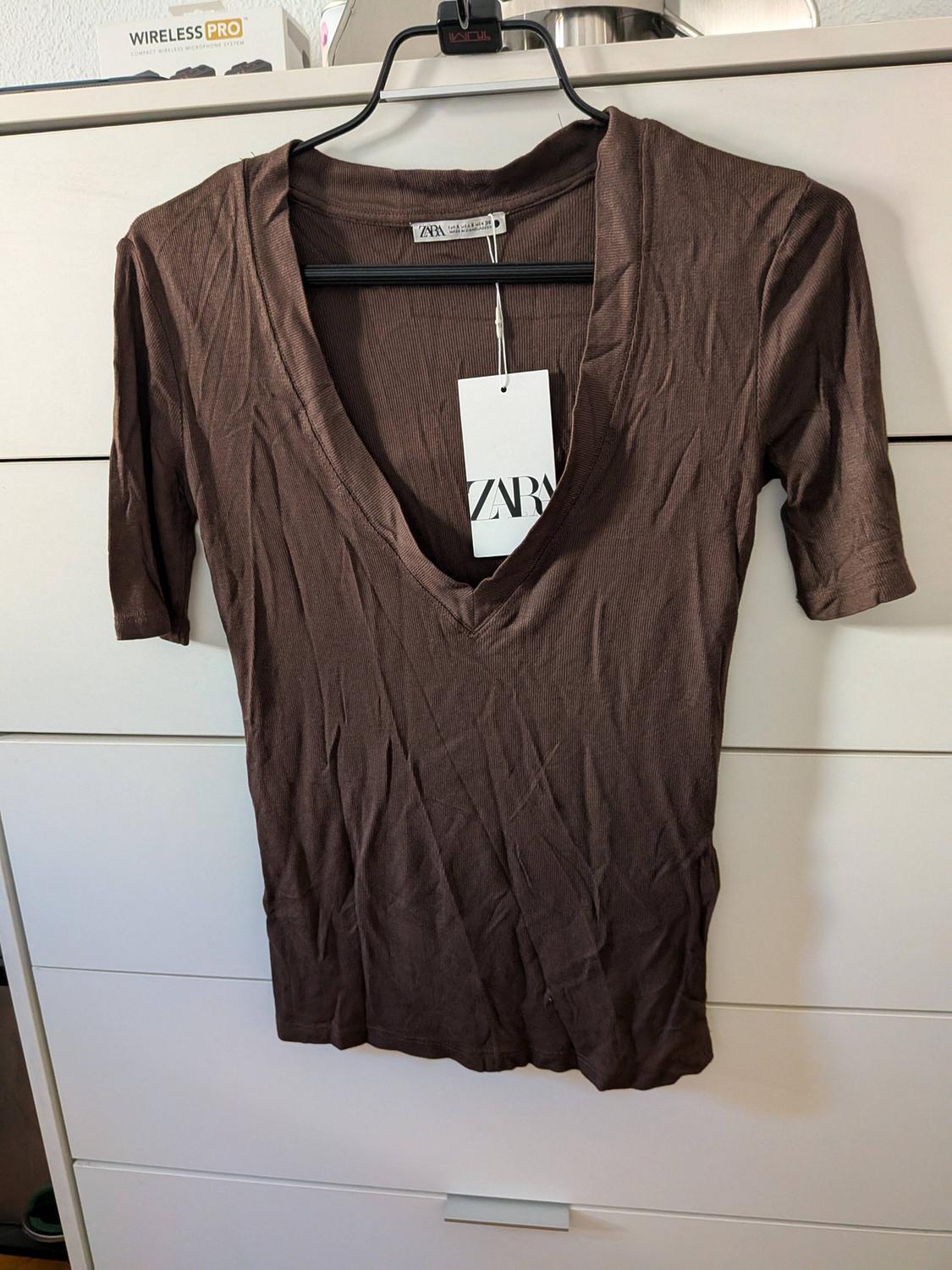 Brown short-sleeved V-neck T-shirt hanging on a hanger in front of white drawers. Texts: ZARA, TUMI, WIRELESS PRO.