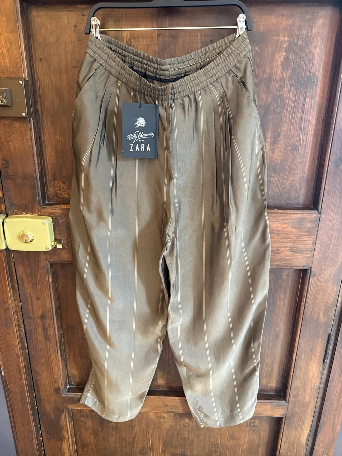 Olive green striped trousers hanging on a rustic wooden door. Label with the text: "Willy Chavarria for ZARA".