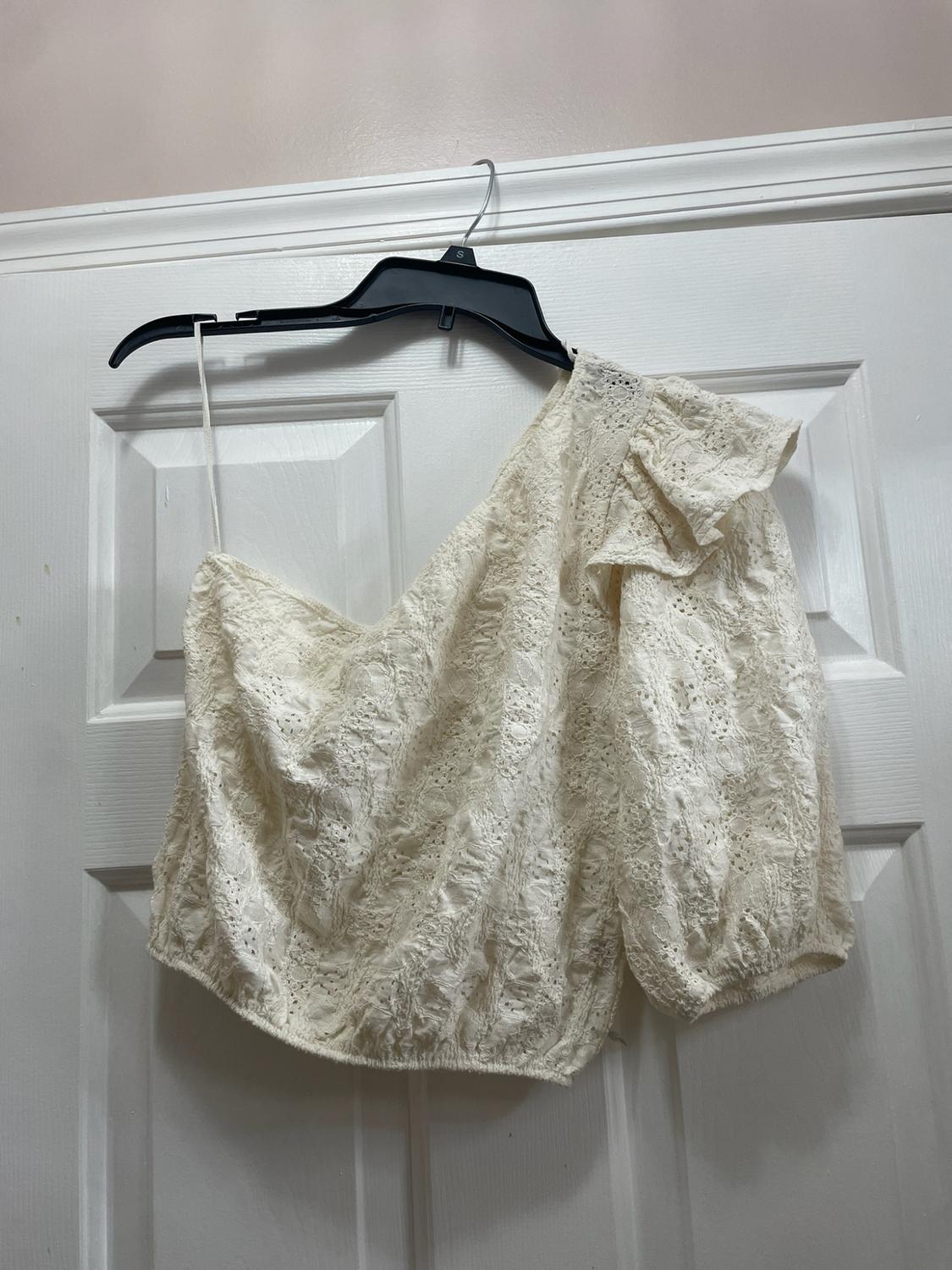 Asymmetrical white lace top with a puff sleeve and thin strap, hanging from a black hanger on a white door.