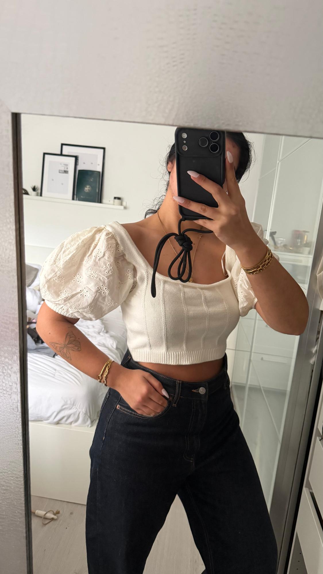 Woman in a white puff-sleeved top and dark jeans, taking a selfie in a mirror. Room with a bed and pictures in the background.