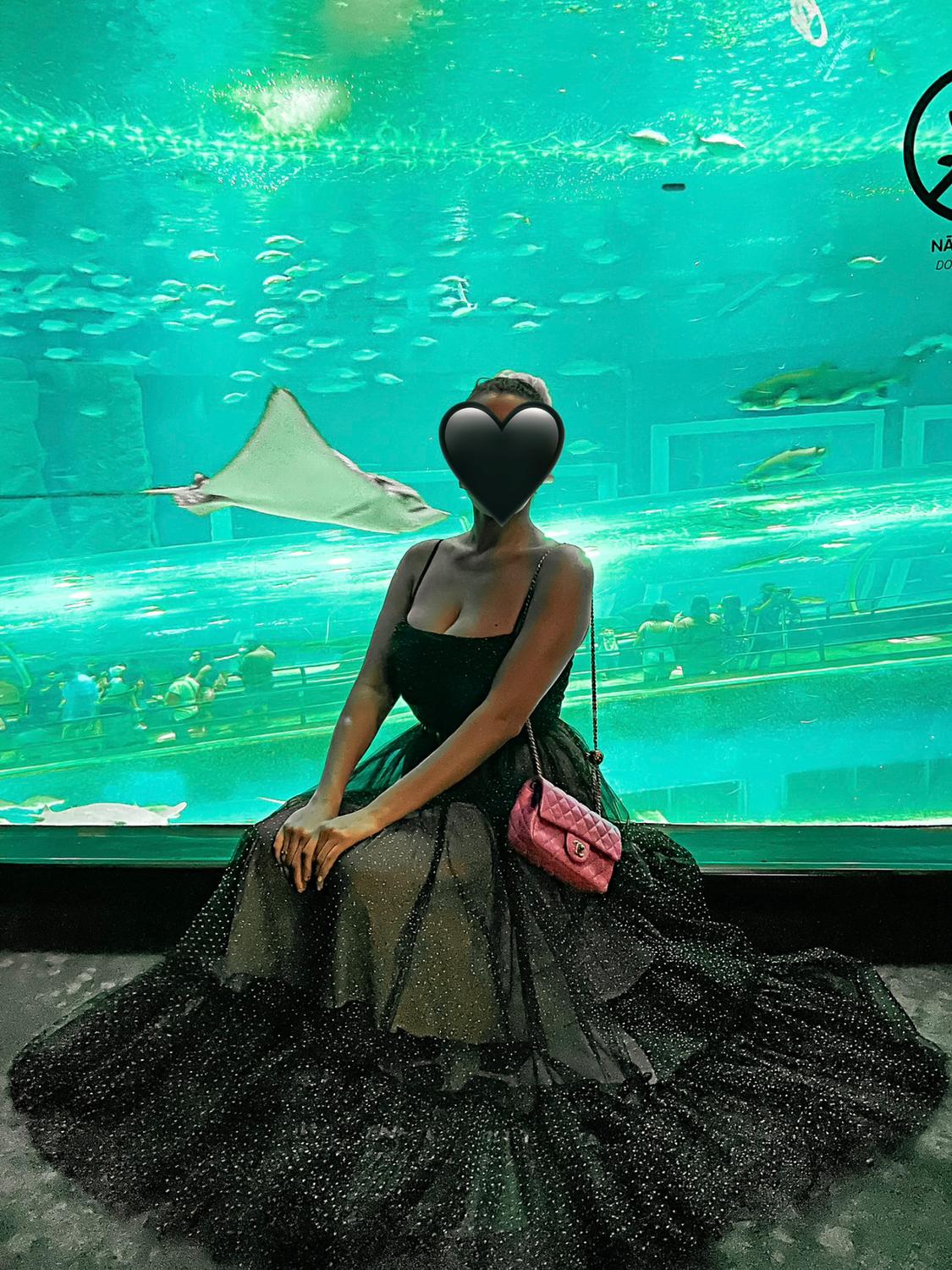 Model in a sparkly black dress and pink handbag, sitting in front of a large aquarium with fish and a ray swimming. Elegant atmosphere.