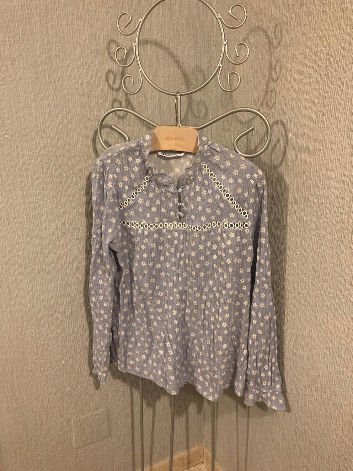 Light blue blouse with a white floral print and openwork details. It hangs from a hanger with the text MANGO on a textured wall.