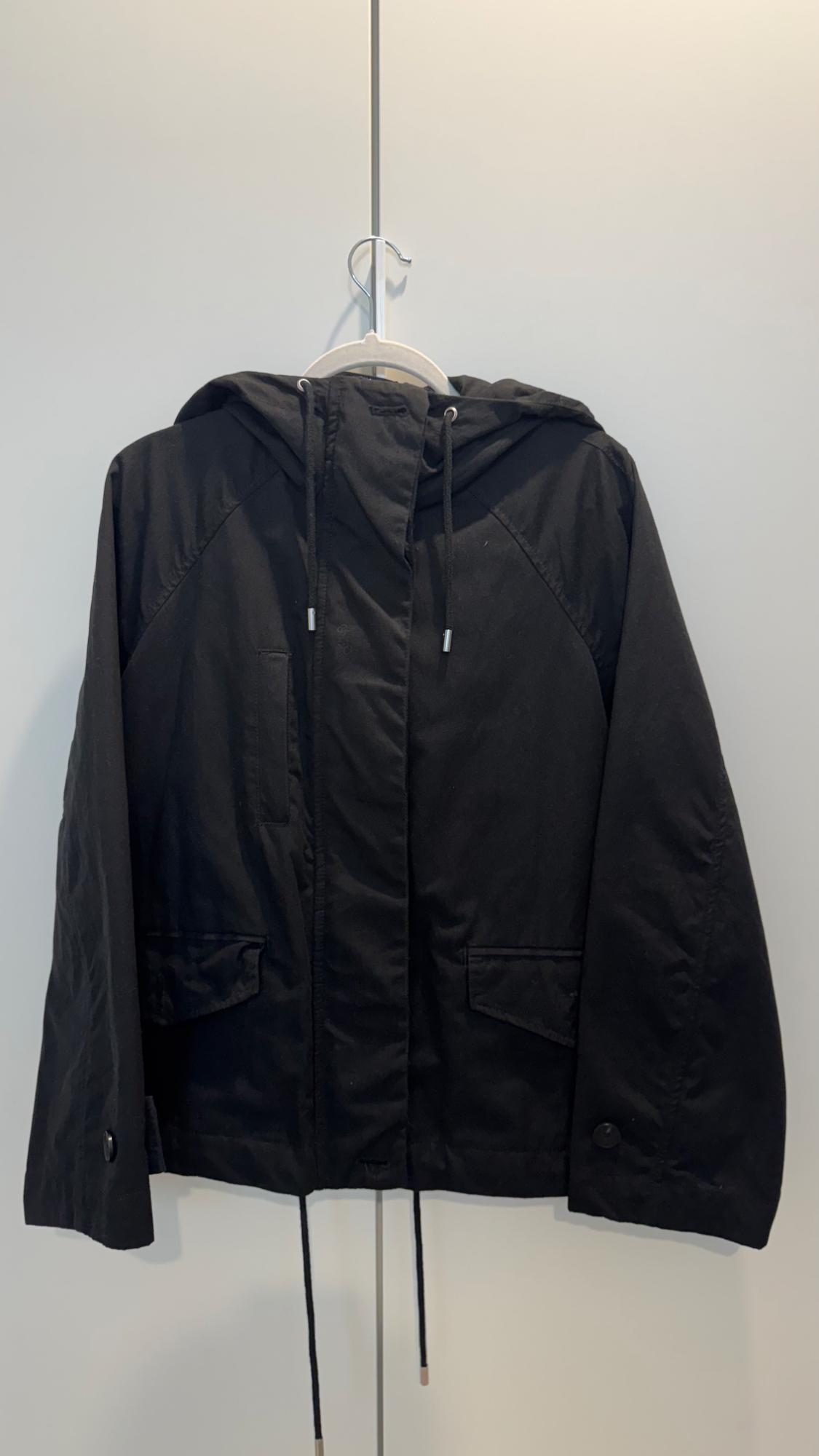 Black hooded jacket with drawstrings, hanging on a white hanger against a light background. Front pockets visible.