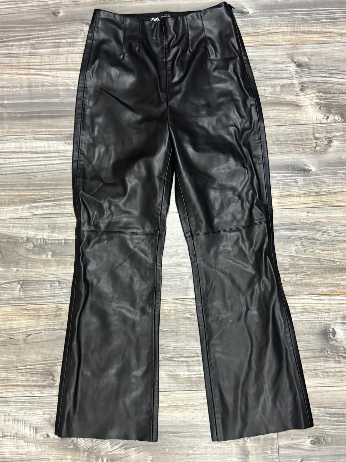 Black faux-leather trousers with a flared hem on a wooden floor. Inner label bearing the text ZARA.