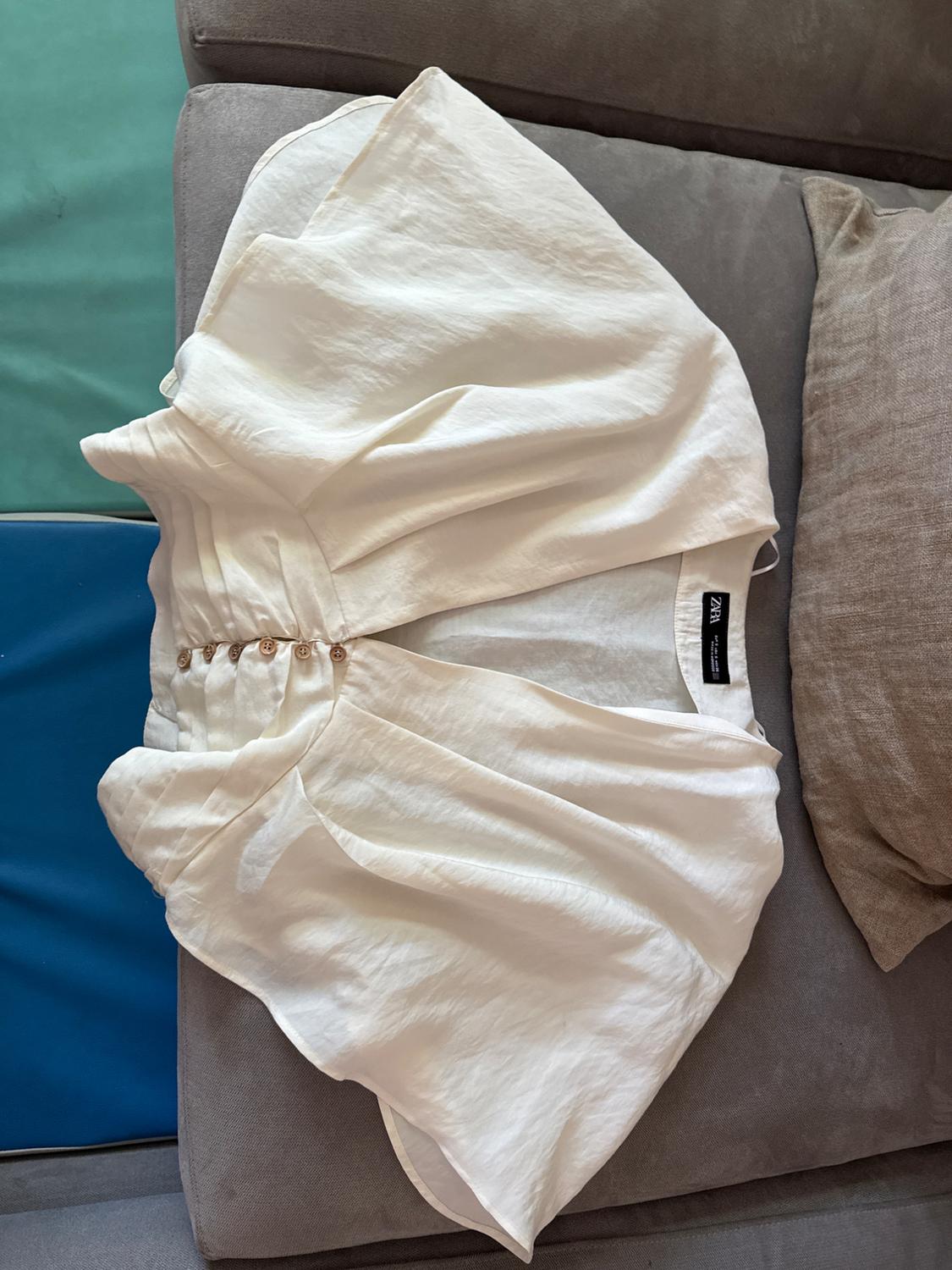 Light-colored white blouse with brown buttons, placed on a gray sofa. Visible "Zara" brand tag.