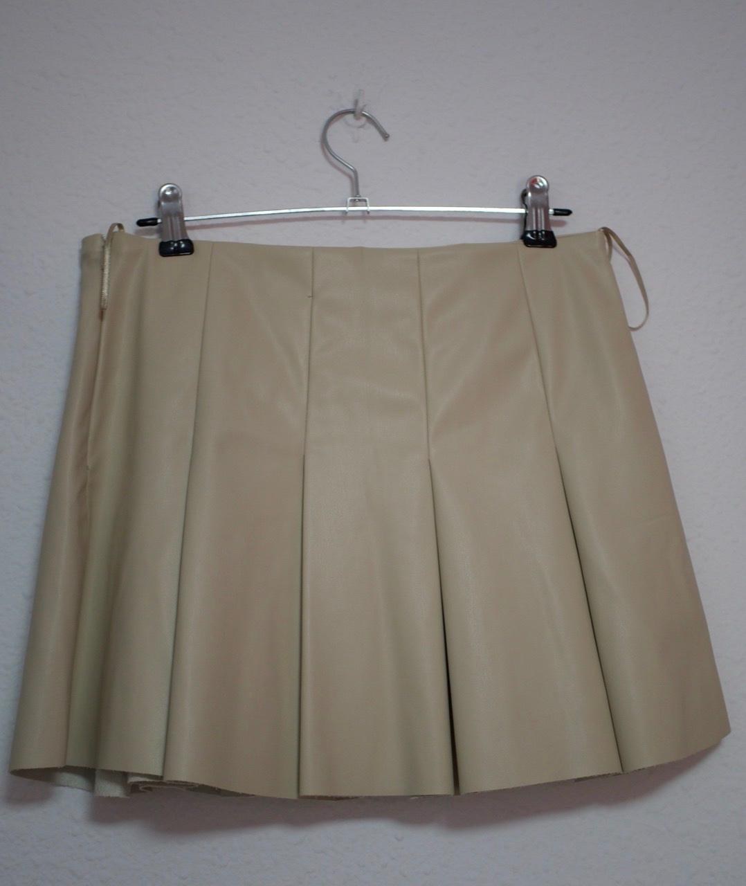 Beige pleated mini skirt hanging on a metal hanger with clips against a white wall background.