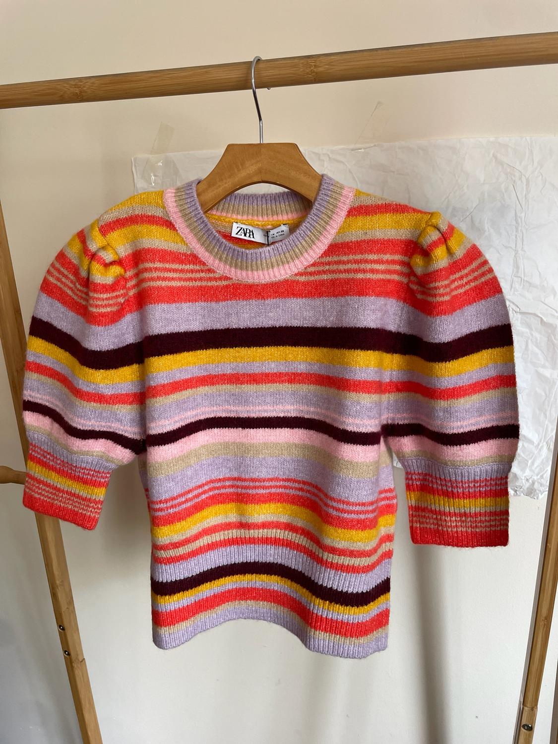 Multicoloured striped jumper with short puff sleeves hanging on a wooden hanger. ZARA label visible at the neckline.