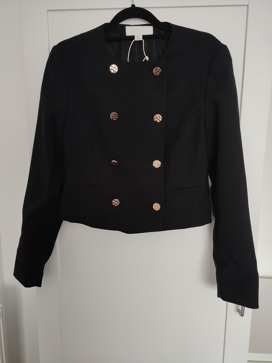 Black long-sleeved, round-neck jacket with double-breasted gold buttons, hanging on a hanger on a white door.