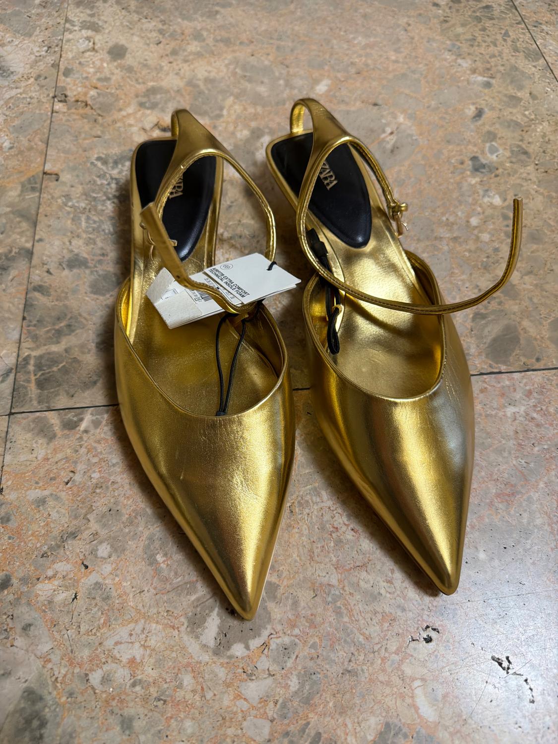 Gold, pointed-toe slingback shoes on a marble floor. Label visible on one of the shoes.
