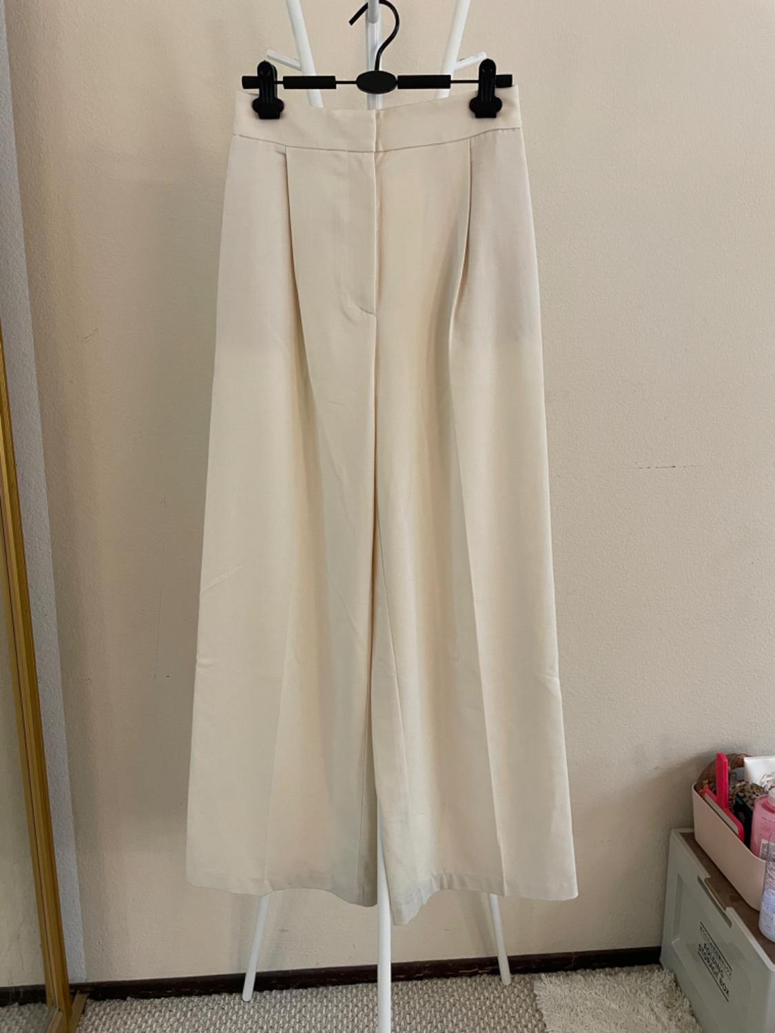 Beige wide-leg pants hanging on a white clothes rack, against a background of light wall and beige carpet.