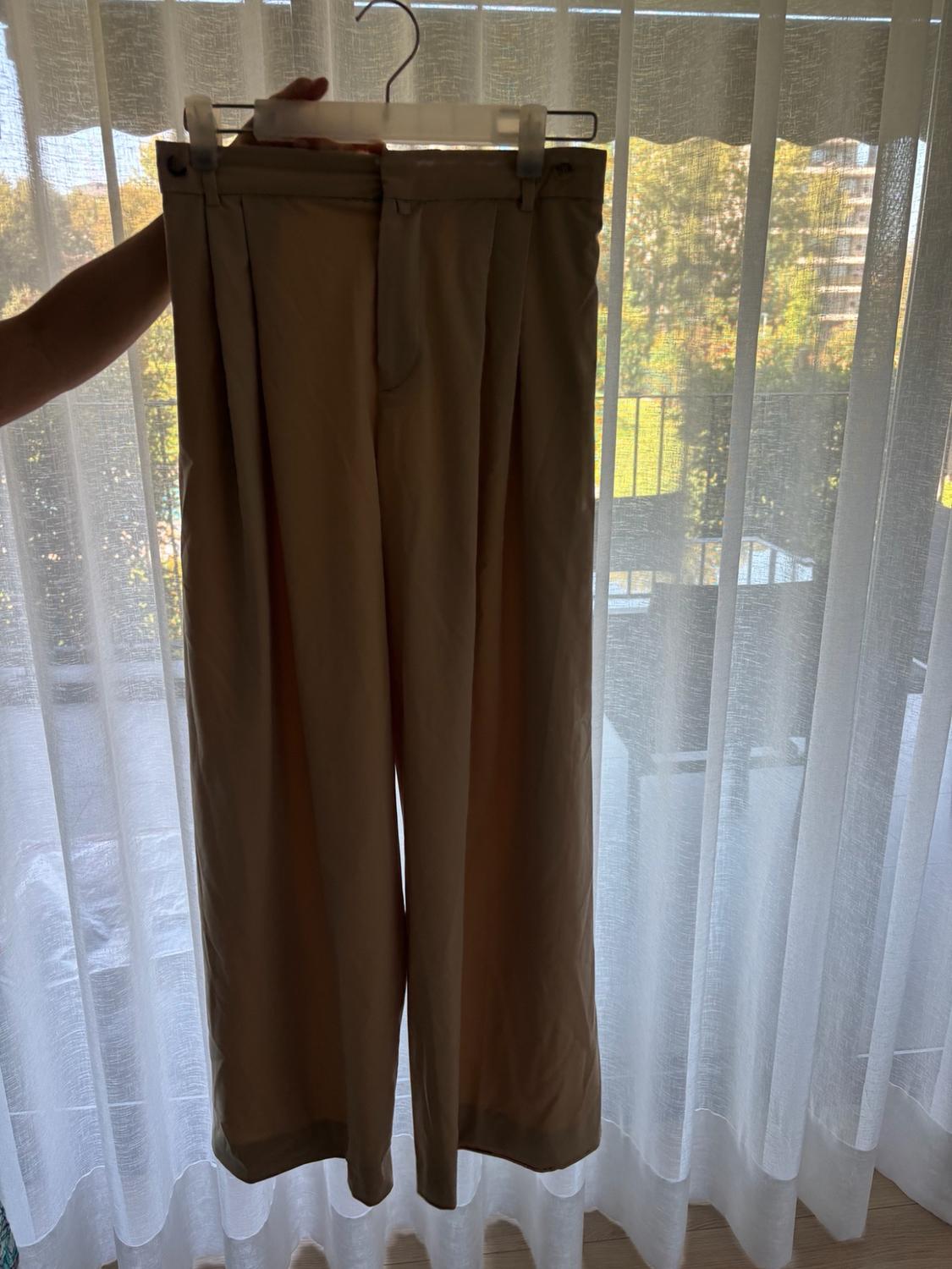 Loose-fitting beige trousers hanging on a hanger held by one hand, positioned in front of a window with natural light and sheer white curtains.
