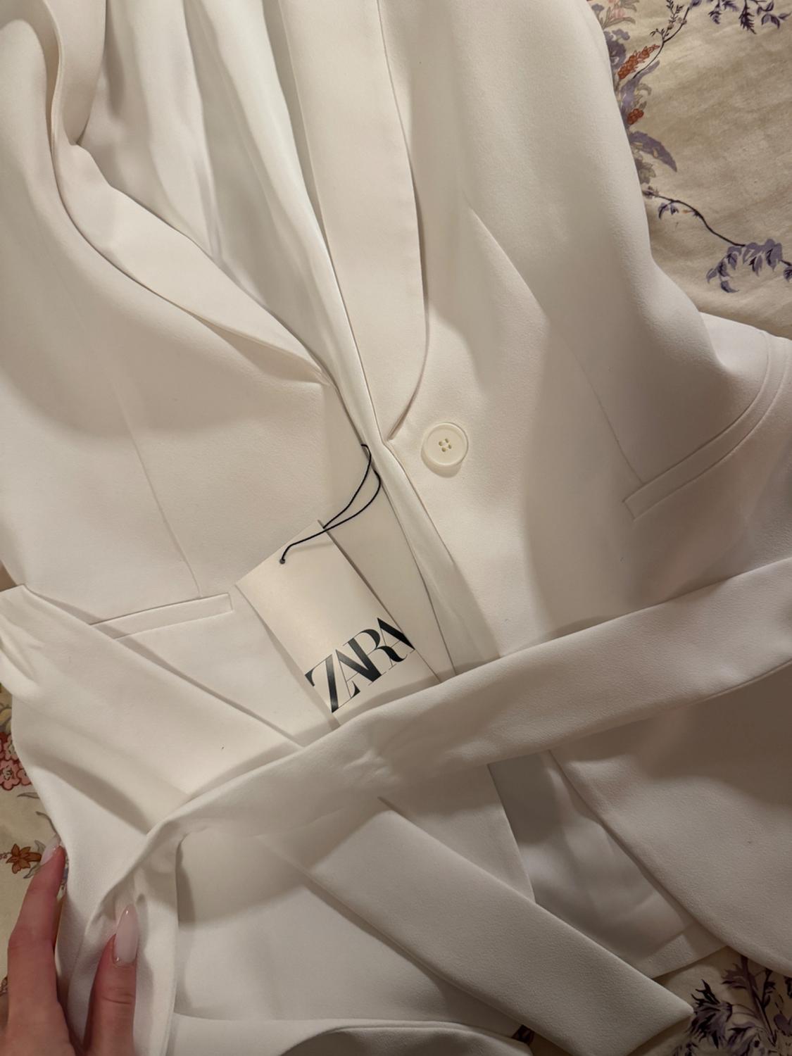 White blazer with a belt over floral fabric, visible tag with "ZARA" text. Hand with long nails holding the garment.
