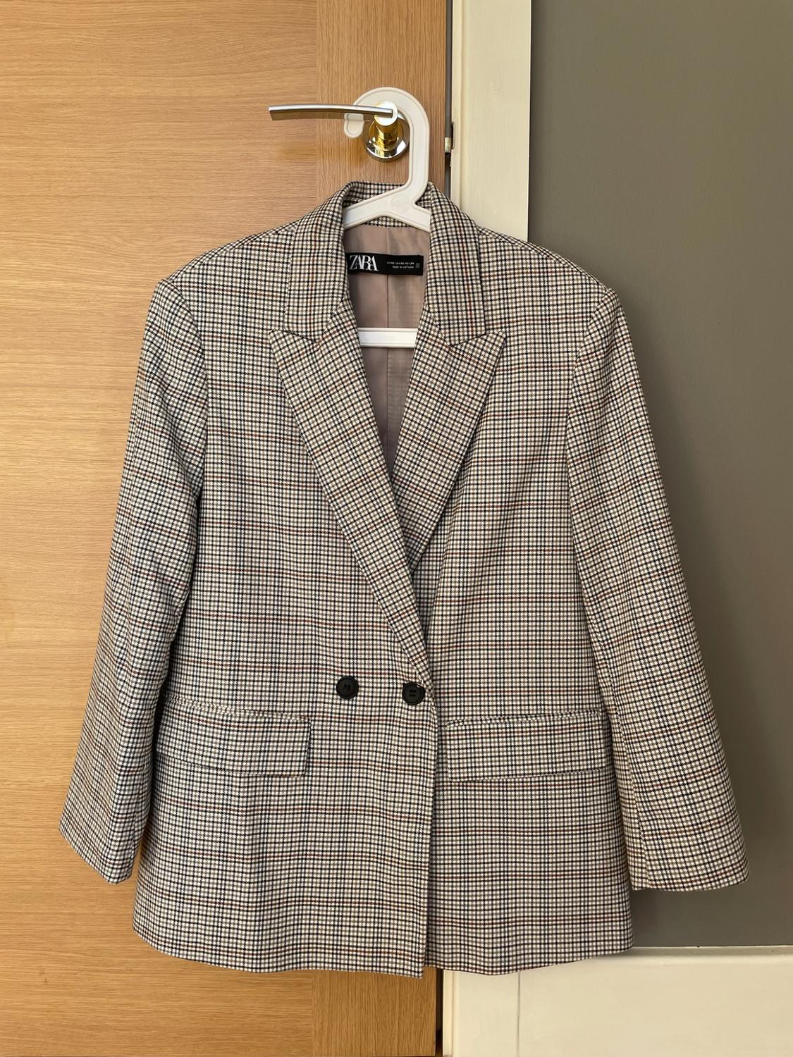 Check blazer with black buttons hanging on a hanger on a wooden door knob. ZARA label.