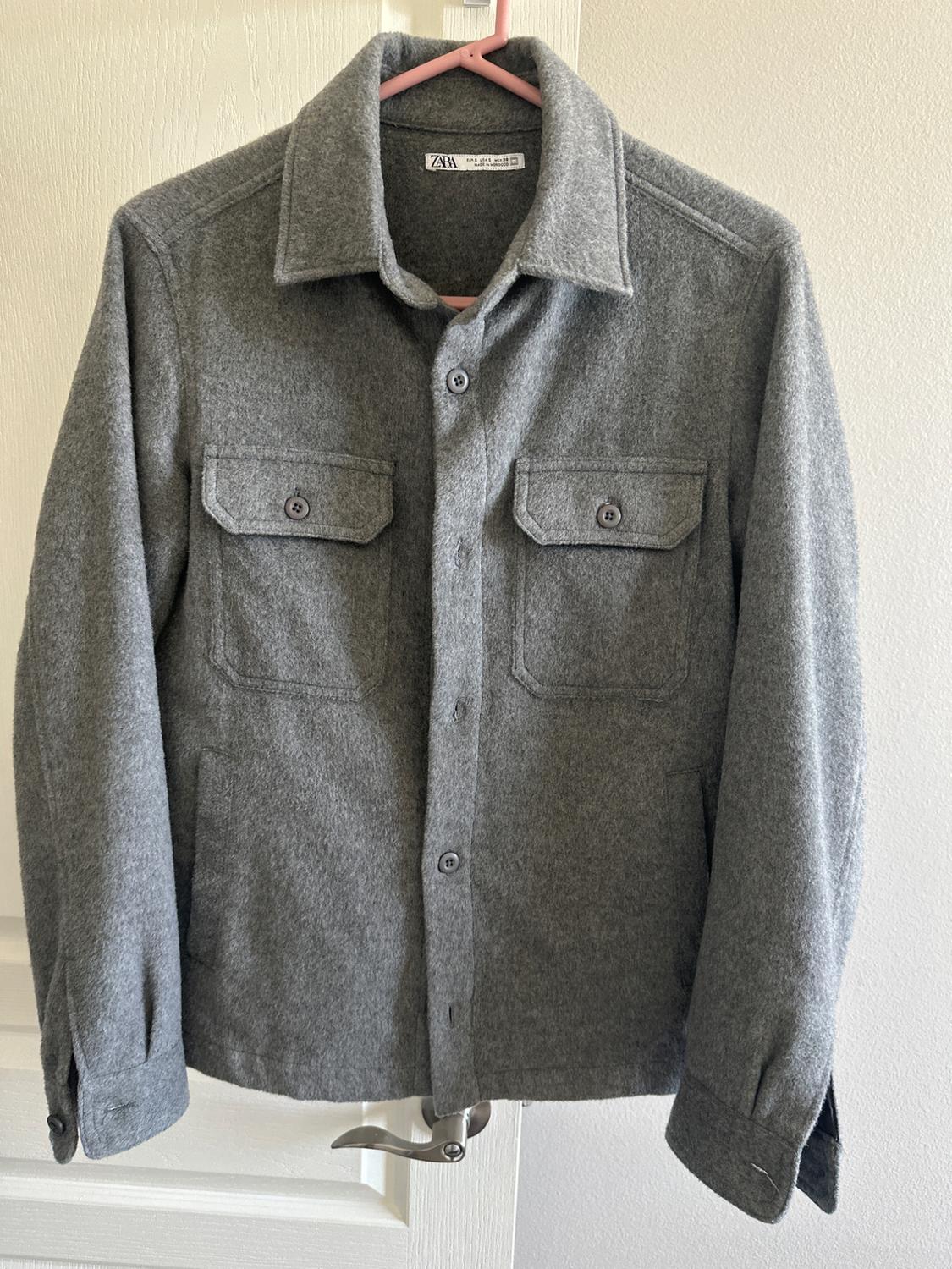 Grey wool jacket hanging on a pink hanger. It has two flap pockets and buttons on the front.