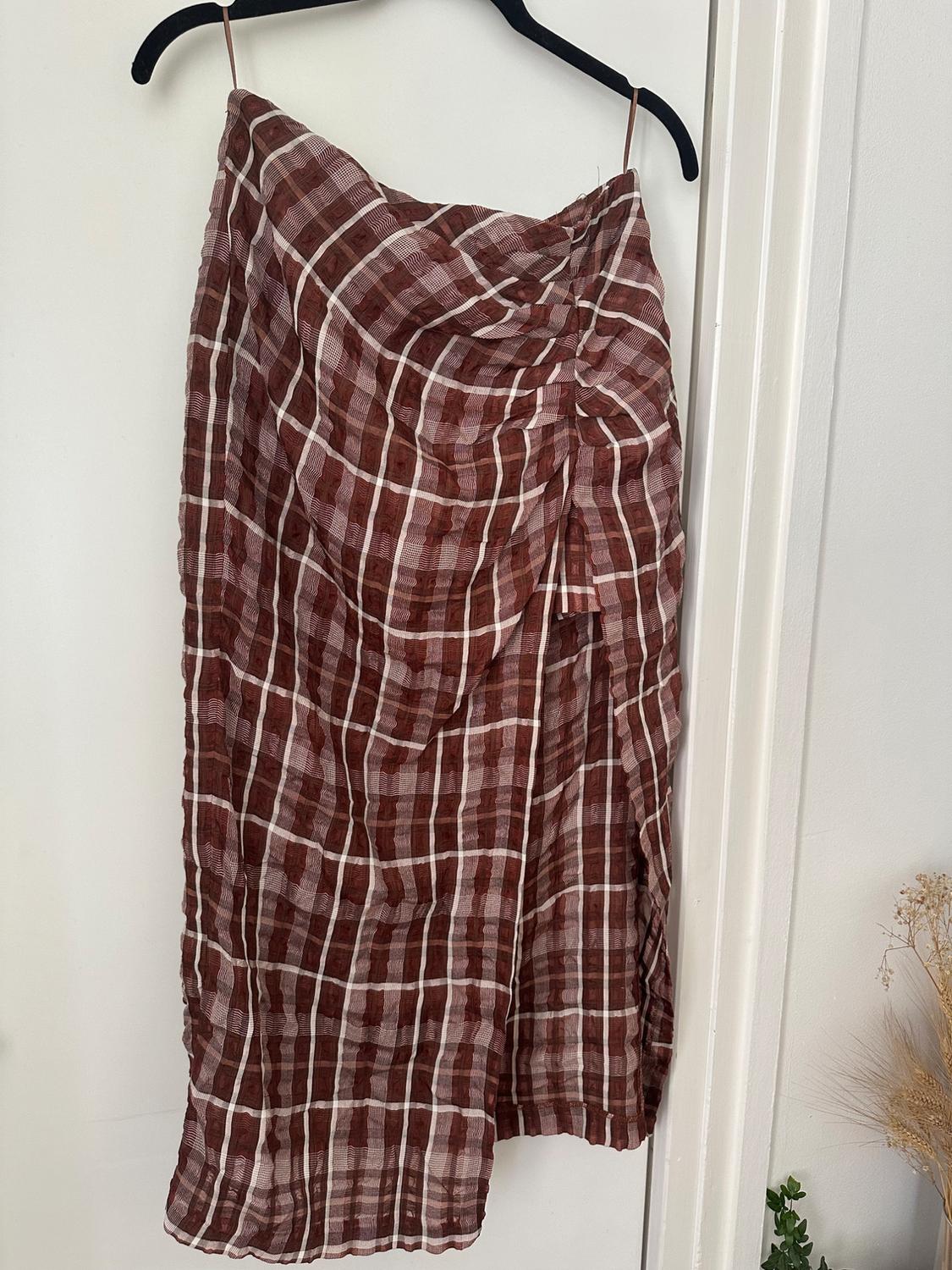 Brown and white checked asymmetric skirt with ruching, hanging on a black hanger over a white door. Dried plants next to it.