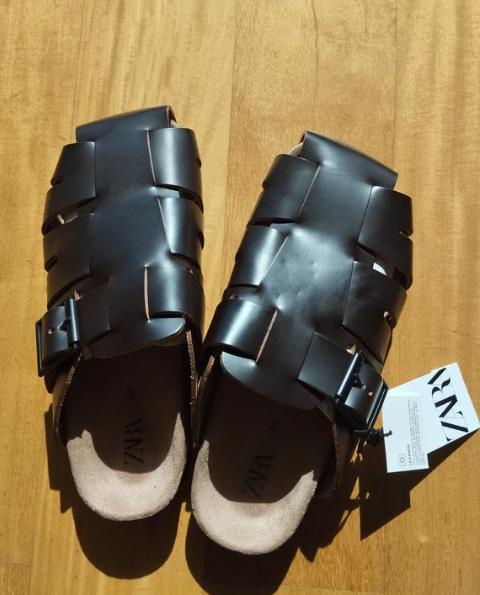 Black leather sandals with an interwoven design and buckles, on a wooden surface. Visible label with the text "ZARA".