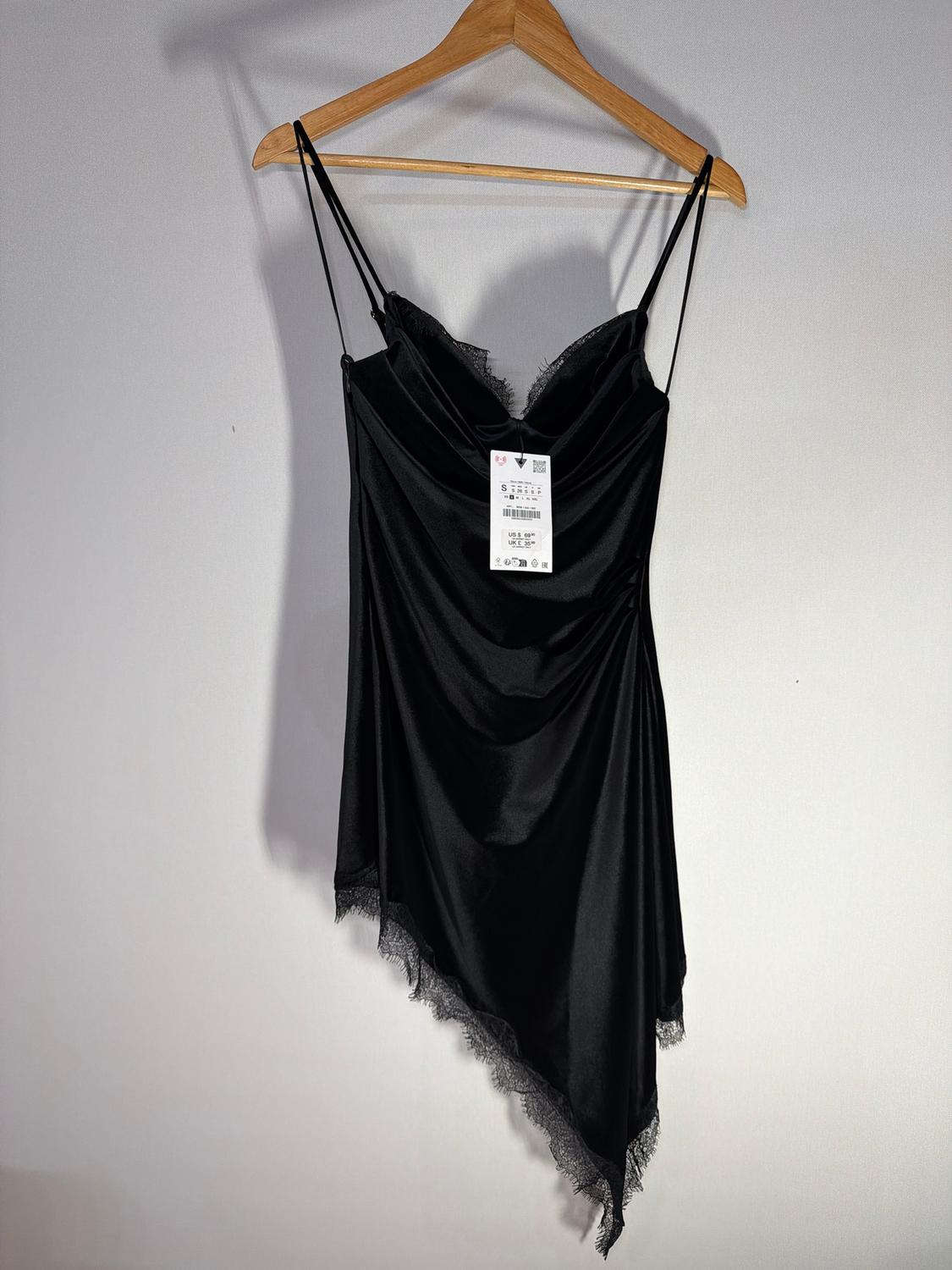 Asymmetrical black dress with spaghetti straps and lace trim, hanging on a wooden hanger against a white background.